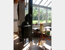 Raum mit einem Kaminofen, großen Fenstern und Dachfenstern, durch die Tageslicht fällt. Im Vordergrund steht ein schwarzer Kamin, während der Hintergrund einen Tisch mit Malutensilien und Blick auf einen Garten zeigt.