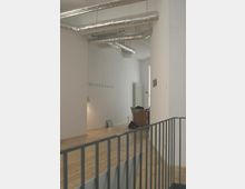 Ein moderner Innenraum mit hellem Holzboden, weißen Wänden und sichtbaren Lüftungsrohren an der Decke. Im Hintergrund befinden sich wenige Möbelstücke und Dekorationen, darunter ein Schreibtisch und eine Lampe.