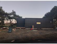 Ein langgestrecktes, modernes Gebäude mit schwarzer Holzfassade und flachem Dach steht auf einem sandigen Grundstück. Davor liegen Baumaterialien und Erdhaufen, umgeben von Kiefern und niedriger Vegetation.