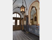 Flur in einem historischen Gebäude mit gewölbter Decke, großen Spiegeln in vergoldeten Rahmen, Marmorsäulen und reich verzierter Wandgestaltung. Eine große hölzerne Doppeltür mit mattem Glas ist im Hintergrund zu sehen, daneben hängt eine antik anmutende Laterne.