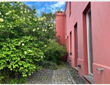 Schmaler gepflasterter Weg entlang einer rosafarbenen Gebäudefassade mit tiefen Fenster- und Türnischen. Links begrenzt dichter, blühender Strauchbewuchs den Weg, mit Kiesstreifen und kleinem Außenwasserhahn an der Wand.