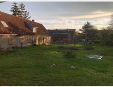 Weitläufiger Hof mit einer großen Wiese, umgeben von Gebäuden mit Ziegeldächern und teils hölzernen Fensterläden; im Hintergrund Bäume und ein blasser Himmel bei Sonnenuntergang.