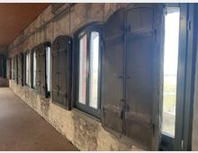 Eine Reihe von Fenstern mit dunkelbraunen Holzklappläden in einer rustikalen Steinmauer, die durch sichtbare Leitungen und eine unbehandelte Holzdecke ergänzt wird. Durch die Fenster ist eine offene Landschaft zu sehen.