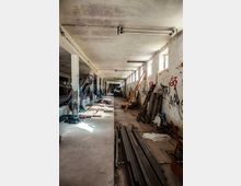 Verlassene Lagerhalle mit einer langen, schmalen Betonstruktur, gestützten Säulen und vergitterten Fenstern an der rechten Wand. Der Raum ist mit Graffiti versehen, teilweise mit Trümmern und Holzstapeln gefüllt, und spärlich beleuchtet durch das einfallende Sonnenlicht.