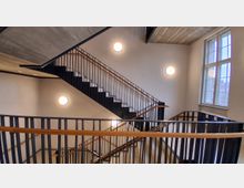 Innenansicht eines modernen Treppenhauses mit mehrläufiger Treppe, schwarzen Metallgeländern und Holzhandläufen. Helle Wände mit runden Wandlampen und ein großes Fenster an der rechten Seite lassen Tageslicht in den Raum.