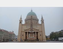 Frontansicht der Nikolaikirche in Potsdam, einem neoklassizistischen Gebäude mit einer mächtigen grünen Kuppel, flankiert von zwei Türmen. Im Vordergrund steht ein Obelisk mit Figuren und Verzierungen, umgeben von einem gepflasterten Platz.