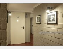 Beleuchteter Flur mit rotem Boden, cremefarbenen Wänden aus Backstein und einer weißen Tür, die mit "Heizung" beschriftet ist. An der Wand hängen schwarz-weiße gerahmte Fotos, beleuchtet von Wandleuchten.