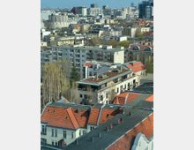 Blick über ein dicht bebautes Stadtviertel mit mehrstöckigen Wohnhäusern, Flachdächern und roten Ziegeldächern. Im Hintergrund sind weitere Wohnblöcke und höhere Büro- bzw. Hochhausgebäude am Stadtrand zu sehen, dazwischen einzelne Baumreihen.