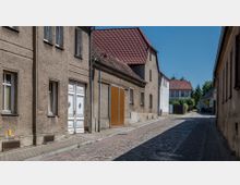 Schmale Kopfsteinpflasterstraße in einem Dorf oder einer Kleinstadt, flankiert von schlichten, verputzten Wohn- und Wirtschaftsgebäuden mit teils roten Ziegeldächern. Im Hintergrund ist ein zweistöckiges Gebäude mit der Aufschrift "Postamt" zu sehen, umgeben von Bäumen.