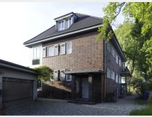 Zweistöckiges Gebäude aus rotem Klinker mit Walmdach und weiß gerahmten Fenstern mit Klappläden. Im Vordergrund ist ein gepflasterter Hof mit angebauter Garage, seitlich umgeben von Bäumen und Grünflächen.