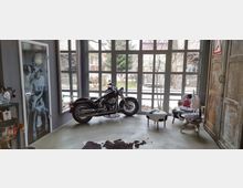 Ein Raum mit großflächigen Fenstern, durch die Tageslicht einfällt, dekoriert mit einer klassischen schwarzen Motorradmaschine im Vordergrund. Links ist eine Tür mit einem Schwarz-Weiß-Bild, rechts stehen Möbelstücke mit Kuhfellmustern und ein Schrank im Vintage-Stil.