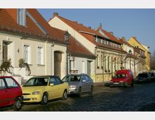 Straße mit gepflastertem Boden und Reihen von Wohnhäusern mit hellen Fassaden und roten Ziegeldächern. Am Straßenrand sind mehrere Autos geparkt, und vereinzelt stehen Bäume sowie Laternen.