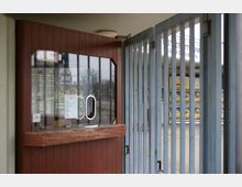 Eine Ticketverkaufsstelle mit hölzerner Verkleidung und Glasscheibe, dahinter ein Metallgittertor. Im Hintergrund sind Plakate und einige Bäume zu sehen.