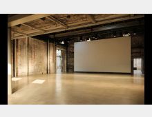 Industriehalle mit Betonboden, freiliegenden Backsteinwänden und sichtbarer Stahlträgerkonstruktion an der Decke. Im Raum steht eine große, weiße Projektionsfläche, die von mehreren Deckenstrahlern beleuchtet wird.