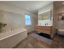 Modernes Badezimmer mit marmorierten Wandfliesen, großer Badewanne links und Doppelwaschtisch mit Holzunterschrank sowie beleuchtetem Spiegelschrank. Rechts ist eine wandhängende Toilette zu sehen; ein Fenster, Einbauspots und dunkle Bodenfliesen prägen den Raum.