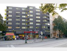Wohngebäude an einer belebten Straße mit dunkelgrauer Fassade und gelb umrandeten Fenstern. Im Erdgeschoss befinden sich ein Supermarkt der Kette "Kaiser's" und ein Imbissstand, umgeben von Bäumen, Gehwegen und einer Straßenlaterne.