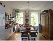 Helle Wohnküche mit großen Fenstern, einem Esstisch mit weißen Stühlen, einer modernen Kücheneinrichtung auf der linken Seite und einem großen Holzschrank auf der rechten Seite. Afrikanische Dekorationen und Pflanzen verleihen dem Raum ein individuelles Flair.