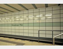 U-Bahn-Station mit einer gefliesten Wand, auf der der Schriftzug „Strausberger Platz“ auf einer grünen Linie steht. Der Boden besteht aus beigefarbenem Stein, und im Vordergrund ist ein Handlauf zu sehen.