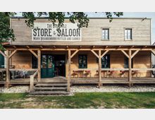 Fassade eines Gebäudes im Westernstil aus Holz mit der Aufschrift "Store & Saloon". Das Gebäude hat eine überdachte Veranda mit Holzbalken, Stühlen und Tischen sowie eine grüne Holztür und Fenster.