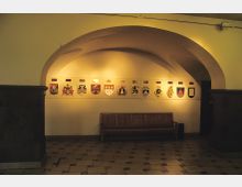 Gewölbter Innenraum mit heller Beleuchtung, an der Wand eine Reihe beleuchteter Wappen auf weißem Hintergrund. Darunter steht eine hölzerne Sitzbank, der Boden ist mit rechteckigen Fliesen verlegt.