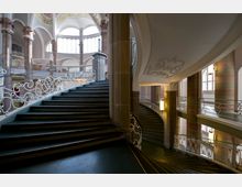 Großzügiges Treppenhaus in einem historischen Gebäude mit kunstvollen, geschwungenen, weißen Geländern aus Metall. Die Architektur wird durch hohe Säulen aus Naturstein, Rundbögen und große Fenster mit diffusem Tageslicht geprägt.