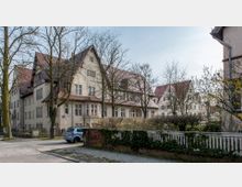 Historisches Gebäude mit mehreren Giebeln und ziegelrotem Dach, umgeben von kahlen Bäumen, einer gepflasterten Straße und einem Zaun mit Efeubewuchs in einem Wohnviertel. Im Vordergrund parkt ein Auto auf der Straße.