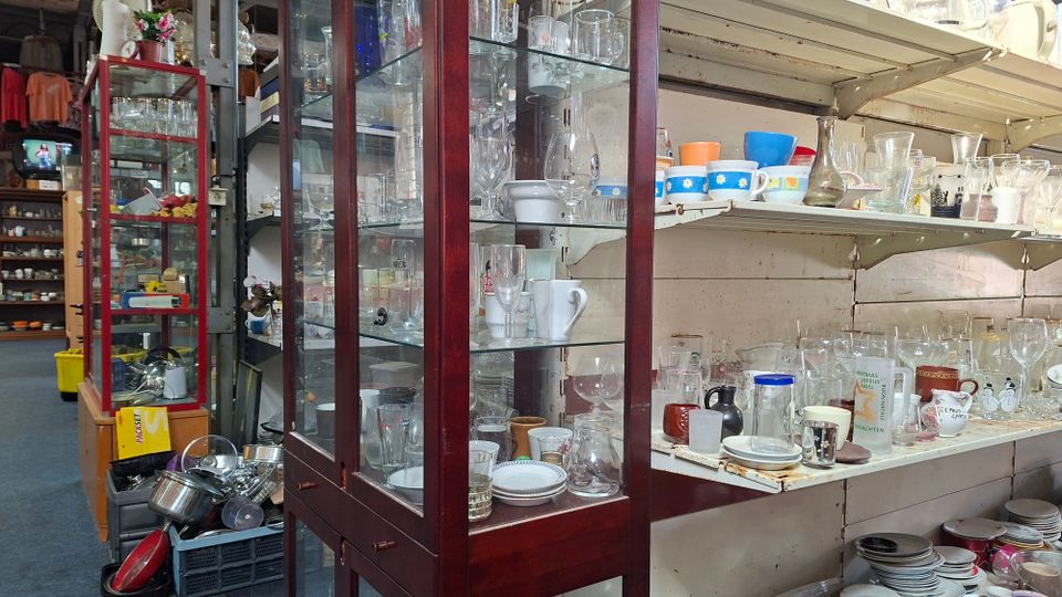 Innenansicht eines Trödelladens mit hohen Glasvitrinen und weißen Wandregalen, dicht gefüllt mit Gläsern, Tassen, Schalen und Geschirr. Zwischen den Regalen stehen Kisten und gestapelte Haushaltswaren, wodurch ein enger, vollgestellter Gang entsteht.