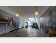 Innenraum eines Museums mit großen Wandbildern, die historische Szenen zeigen. In der Mitte des Raums befindet sich eine kleine Telefonzelle, dahinter eine breite Treppe mit Geländern, die zu einem Fenster führt, das Tageslicht hereinlässt. Drei hängende Lampen beleuchten den Raum.