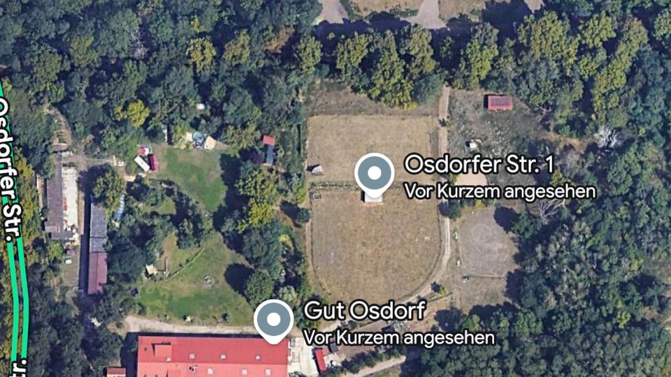 Satellitenansicht eines ländlichen Gebiets mit einem großen Waldstück, einer gerodeten Lichtung mit Wegen sowie einem Gutshof mit langem roten Gebäude und mehreren Nebengebäuden. Am unteren Bildrand sind Felder und ein gerader Weg bzw. eine Trasse zu erkennen.