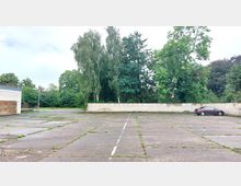 Freifläche mit betonierten Parkplätzen, umgeben von einer niedrigen weißen Ziegelmauer und dichtem Baumbestand im Hintergrund; ein einzelnes geparktes Auto steht rechts im Bild.