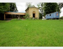 Große Wiese mit einem kleinen Backsteingebäude im klassischen Stil und einem flachen Anbau aus Holz und Glas links. Rechts steht ein blauer Wohnwagen neben einem geparkten Auto, umgeben von Bäumen.