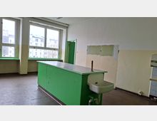 Leerer, schlicht eingerichteter Innenraum mit großen Fenstern und einem langen grünen Arbeitstisch mit integriertem Waschbecken und Wasseranschluss. Helle Wände mit abgenutzten Stellen und eine grün gestrichene Tür prägen den Raum.