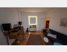 Wohnzimmer mit Holzboden, einer blauen Eckcouch, einem Teppich und einem kleinen weißen Beistelltisch. An der linken Wand befindet sich eine Holzkommode mit DJ-Ausrüstung und Lautsprechern, während an der gegenüberliegenden Wand eine leuchtende quadratische LED-Dekoration über einer weißen Kommode hängt. Luftballons liegen verstreut auf dem Boden.