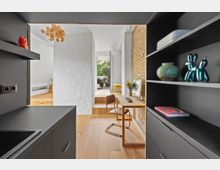 Moderne Wohnraumgestaltung mit Blick aus einer kleinen Küche in ein helles Arbeits- und Wohnzimmer. Rechts ist eine Backsteinwand mit Regalen und Dekorationselementen, links ein schwarzer Küchenschrank, und im Hintergrund ein Holzschreibtisch vor einer Glastür, die auf eine Terrasse mit Pflanzen führt.