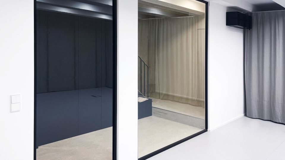 Minimalistischer Innenraum mit weißen Wänden, großen Glaswänden in schwarzem Rahmen und hellgrauem Vorhang. Im Hintergrund ist ein Treppenbereich mit Sichtbeton und leicht industriellem Design sichtbar.