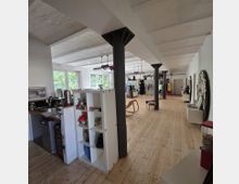 Weitläufiger Raum mit Holzboden und weiß gestrichenen Wänden, durchzogen von schwarzen Stahlträgern. Links befindet sich eine offene Küche, während der Rest des Raums mit Regalen, Sitzmöbeln und Dekorationen ausgestattet ist; große Fenster lassen Tageslicht herein.