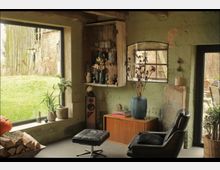 Gemütlicher Wohnraum mit rustikaler Einrichtung, einschließlich grüner Wände, einem großen Fenster mit Blick auf eine Wiese, einem Ledersessel mit Fußhocker und Holzdekor. Neben einer Vitrine befinden sich eine Vase mit getrockneten Blumen und ein Holzschrank mit Büchern und Dekorationsgegenständen.