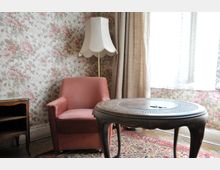 Wohnzimmer in einem retrogestalteten Stil mit einer floral gemusterten Tapete, einem rosa Sessel, einem hölzernen Beistelltisch und einer Stehlampe mit einem geschwungenen weißen Lampenschirm. Im Hintergrund sind lichtdurchlässige Vorhänge vor einem Fenster zu sehen.