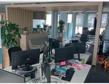 Ein modernes Büro mit mehreren Schreibtischen, Computermonitoren und Bürostühlen. Im Hintergrund befinden sich große Fenster mit Rollläden, Pflanzen und eine minimalistisch gestaltete Sitzecke mit einem Sofa und einem kleinen Tisch.