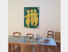 Ein Esszimmer mit Holzboden, einem großen Glastisch und unterschiedlichen Holzstühlen. An der weißen Wand hängt ein buntes Gemälde mit drei gelben Figuren auf grünem Hintergrund.