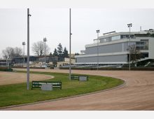 Eine Pferderennbahn mit einer sandigen, ovalen Rennstrecke, umgeben von Rasen. Im Hintergrund befinden sich moderne Tribünengebäude mit großen verglasten Fassaden und Flutlichtmasten, sowie einige Bäume.