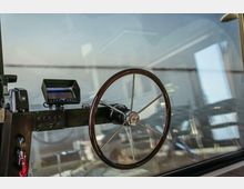 Eine Nahaufnahme eines Bootssteuerrads aus Holz und Metall, eingebaut in eine Cockpit-Konsole mit einem kleinen Bildschirm und weiteren Bedienelementen. Im Hintergrund ist durch die Scheibe verschwommen Wasser und ein helles Tageslicht zu erkennen.