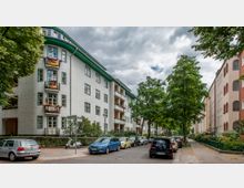 Wohnstraße mit mehrstöckigen, hell verputzten Wohnhäusern und einem grünen Dachrand, mehreren Balkonen und dekorativen Details. Am Straßenrand stehen parkende Autos und vereinzelte Bäume, die den Gehweg säumen.