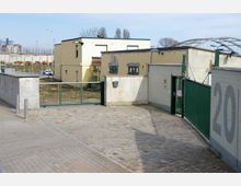 Eingezäunter Hofbereich mit gepflastertem Boden, umgeben von hellgelben und grauen Gebäuden mit flachen Dächern. Im Hintergrund sind ein Geländer, eine Brücke und industrielle Strukturen zu sehen.