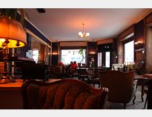 Gemütliches Café mit Vintage-Einrichtung, darunter antike Polstermöbel, eine Tischlampe und dunkle Holzelemente an Fenstern und Wänden. Im Hintergrund sitzen einige Gäste an Tischen vor großen Fenstern, die Tageslicht hereinlassen.