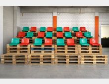 Provisorische Tribüne in einer Industriehalle: bunte Kunststoffsitze in Rot und Grün stehen in mehreren Reihen auf gestapelten Holzpaletten. Im Hintergrund eine weiße Ziegelwand mit orangefarbenen Stützen und grauer Beton­decke, davor ein gefliester Boden.
