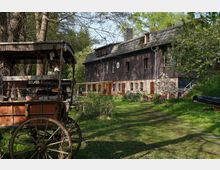 Ein rustikales Wohnhaus mit dunkler Fassade und Ziegelsockel steht inmitten einer grünen, blühenden Gartenlandschaft. Im Vordergrund befindet sich ein hölzerner, alter Wagen, umgeben von Bäumen und grasbewachsenem Gelände.