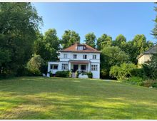 Zweistöckiges weißes Haus mit rotem Dach und zahlreichen Fenstern, umgeben von einem großen Rasen und dichten Bäumen im Garten. Die Umgebung wirkt ländlich-grün mit mehreren Sitzgelegenheiten und Pflanzen im Vordergrund.