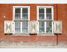 Ziegelsteinfassade eines Gebäudes mit zwei Doppelfenstern, umrahmt von verwitterten weißen Holzläden. Hinter den Fenstern sind weiße Vorhänge und Pflanzendekorationen sichtbar.