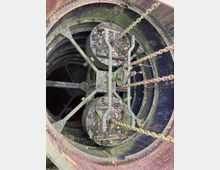 Blick in den Innenraum eines alten, moosbedeckten Turms oder Schachts mit großen Metallrädern und angehängten Ketten. Die Oberflächen sind von Blättern und Verschmutzungen bedeckt.